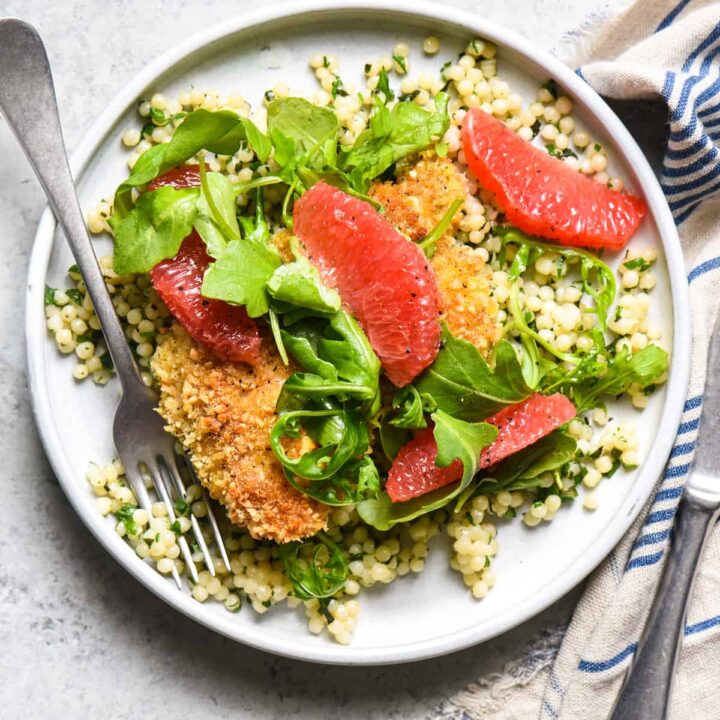 A small white plate of baked almond chicken with couscous, salad and grapefruit on a gray surface.