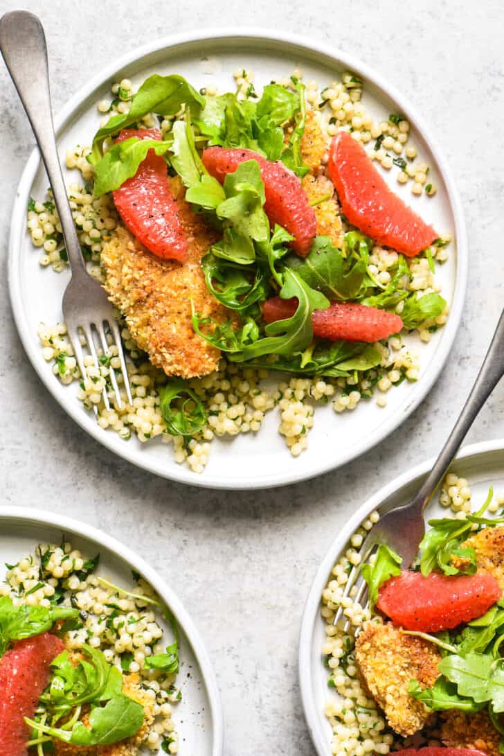 Three small gray plates topped with almond crusted chicken, Israeli couscous, arugula and grapefruit segments.