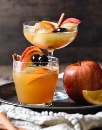 Apple cider old fashioned cocktails in two different glasses (1 tall, 1 short), on a tray with a whole apple.