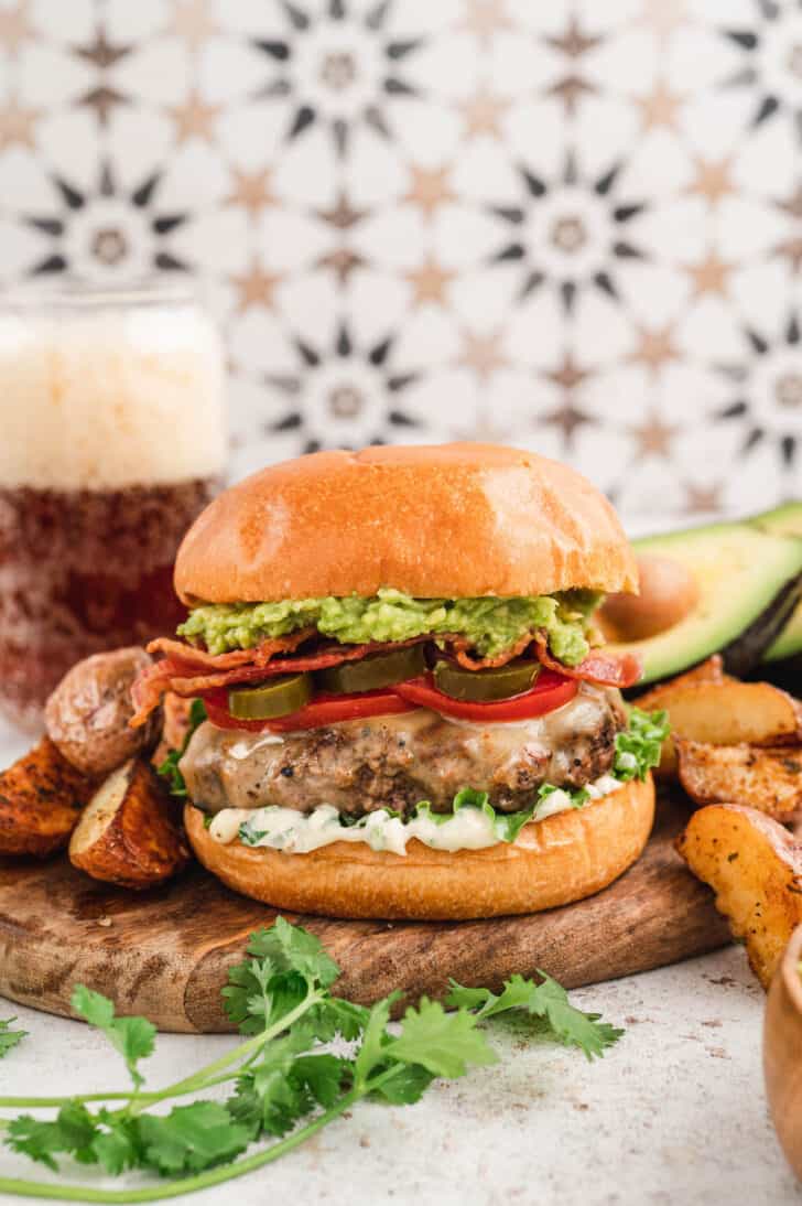 An avocado bacon burger on a brioche bun topped with lettuce, tomato and pickled jalapeno. The burger is on wooden cutting board in front of a patterned background. There are potato wedges in the foreground.