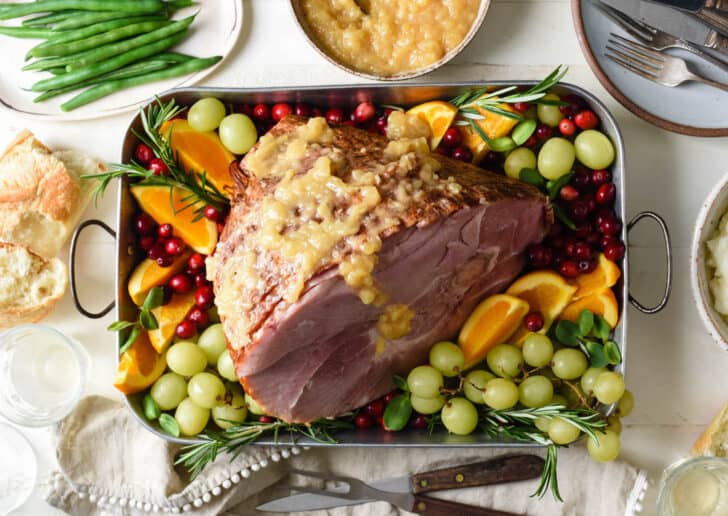 Metal roasting pan with spiral sliced ham topped with pineapple sauce, and surrounded by fruit garnishes.