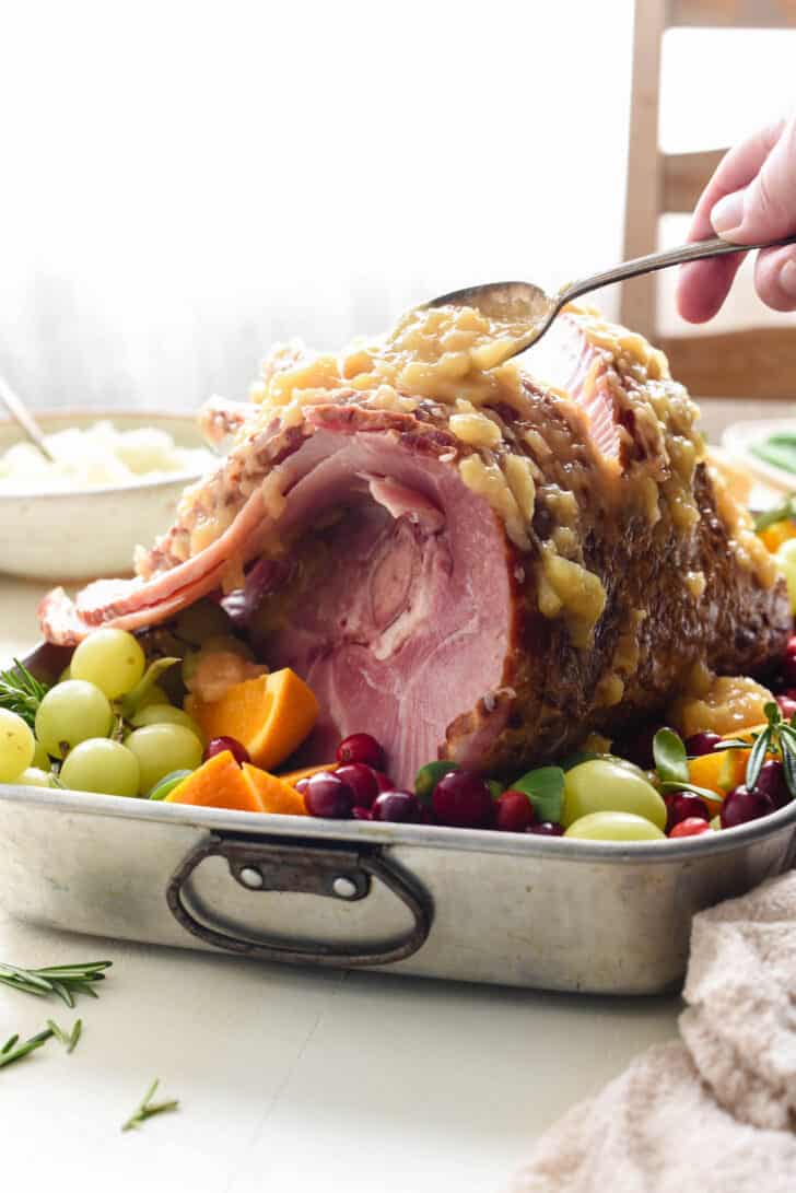 Metal roasting pan with spiral sliced ham surrounded by fruit garnishes, with woman's hand spooning pineapple sauce over ham.