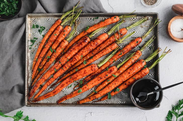 A textured baking pan topped with balsamic glazed carrots with their green tops still on, with a small bowl of balsamic glaze on the pan.