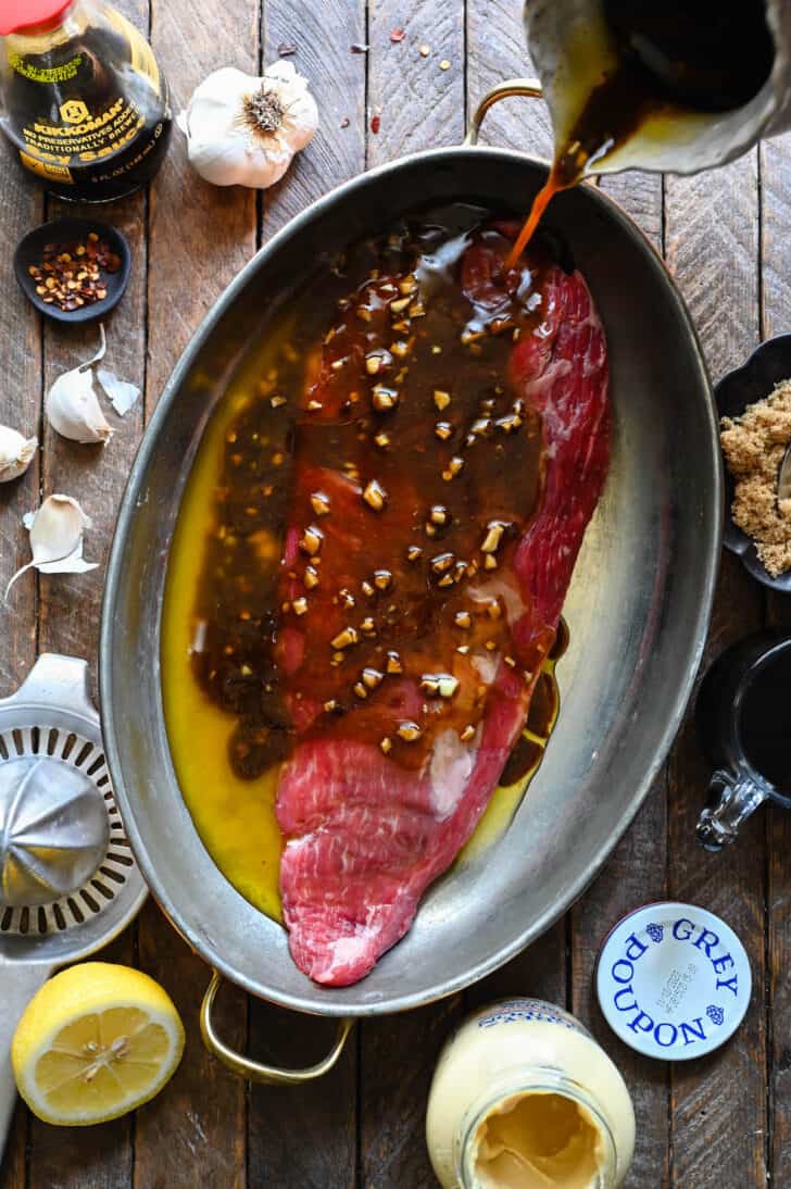 A piece of beef in a oval pan, with a hand pouring a dark liquid mixture with flecks of garlic in it over the meat.