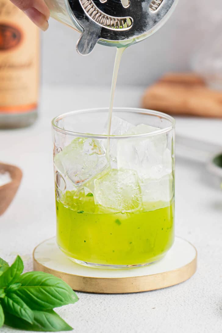 A cocktail strainer pouring a bright green liquid over ice cubes in a short glass.