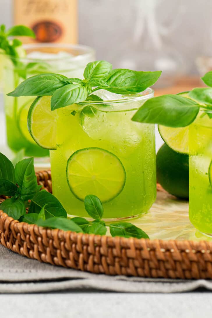 Green hued basil cocktails garnished with lemon on a wicker tray.