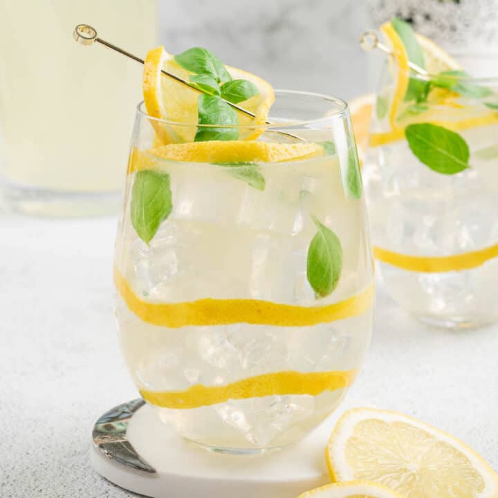 A glass filled with ice and basil lemonade, garnished with lemon peels and fresh basil leaves.