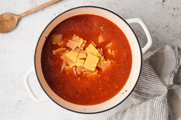 A white Dutch oven filled with a red soup mixture, with broken lasagna noodles on top.