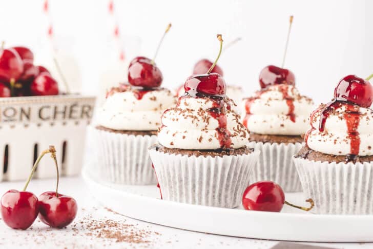 A white plate of Black Forest cupcakes, which are chocolate cupcakes topped with whipped cream, cherries and chocolate shavings.