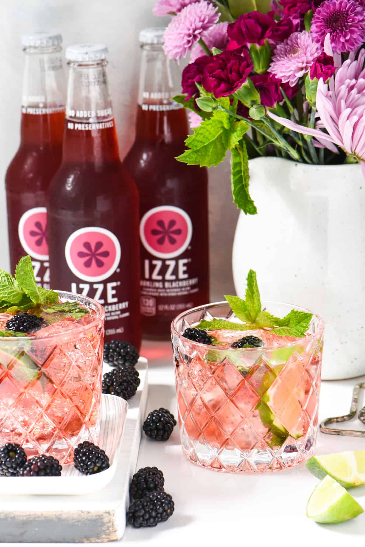 Two double old fashioned glasses filled with a pink drink, garnished with mint. Blackberry soda bottle and a vase of purple flowers are in the background.