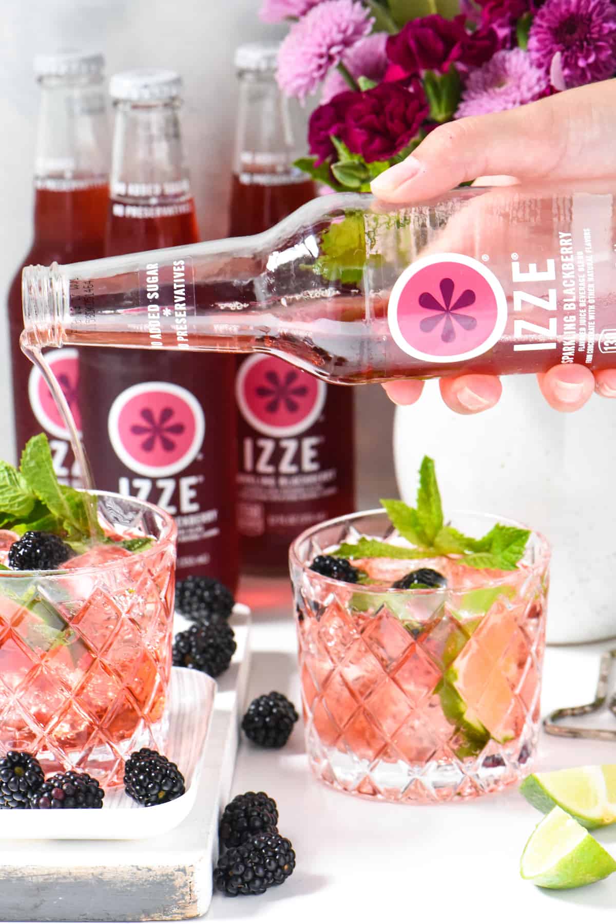 Two small glasses of a pink cocktail garnished with mint and blackberries, with a hand pouring blackberry soda into one of the glasses.