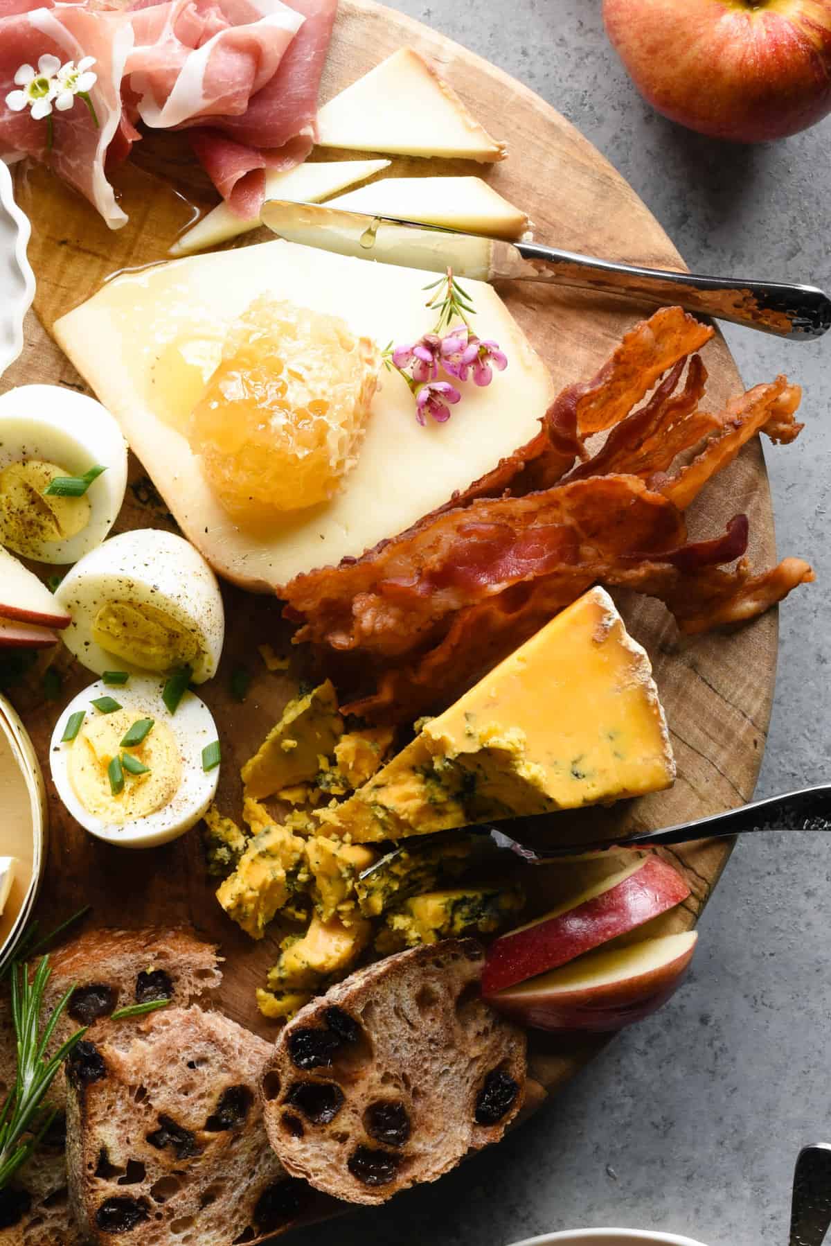 Round wooden cutting board topped with cheese, bacon, honeycomb and hard boiled eggs.