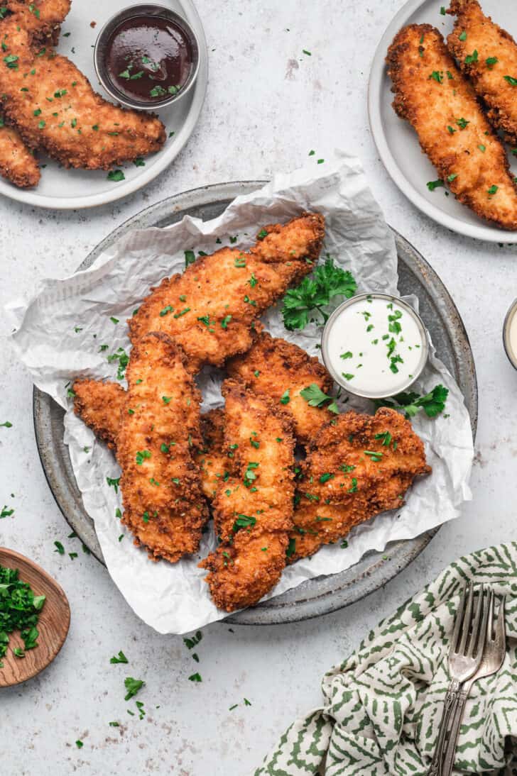 Fried chicken fingers on a parchment-lined plate with a side of ranch dressing, sprinkled with parsley.