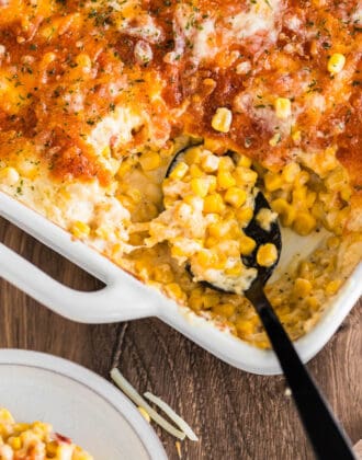 A white baking dish full of cheesy corn casserole with a black spoon taking a scoop out.