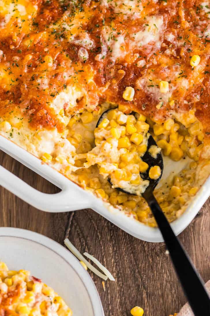 A white baking dish full of cheesy corn casserole with a black spoon taking a scoop out.