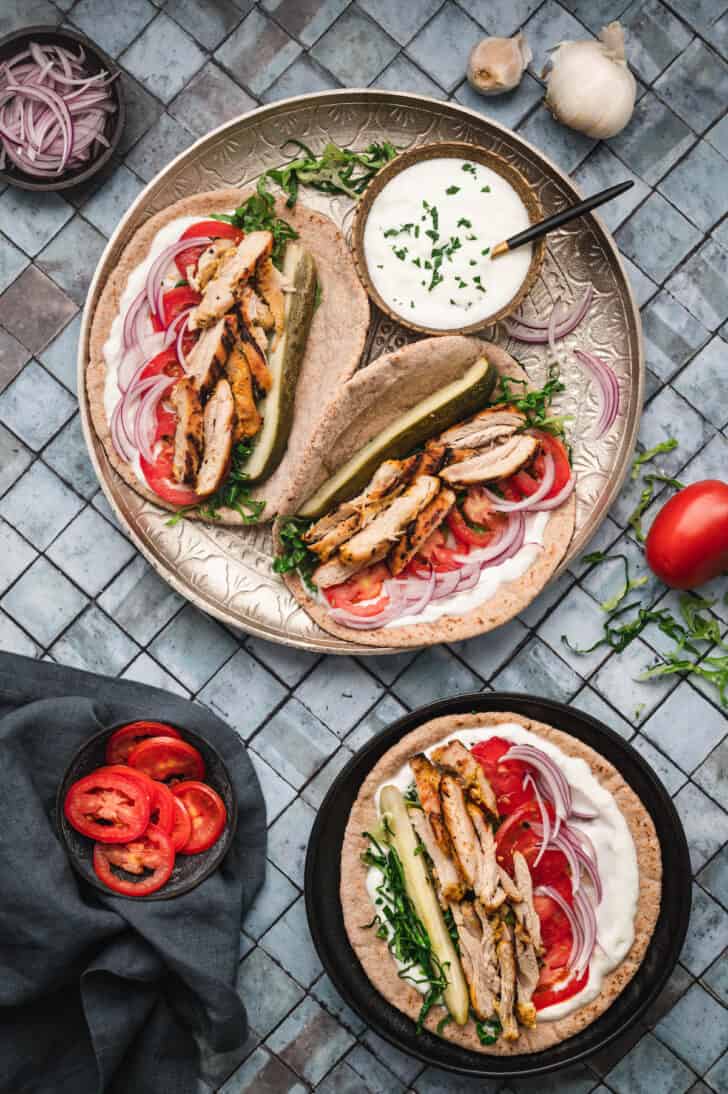 Two wrap sandwiches on a patterned tray with a a bowl of yogurt sauce, with a third pita on a smaller black plate.