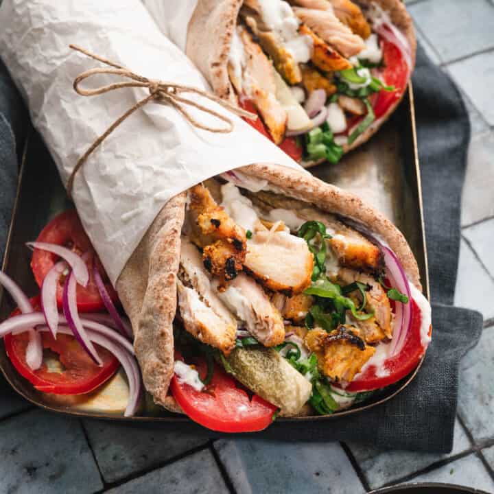 Two grilled chicken shawarma wraps, wrapped in white paper, on a gold platter on a tiled surface.