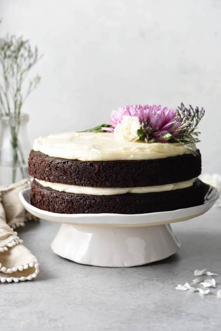 A two layer chocolate cake with cream cheese frosting on a white cake stand, decorated with pink and white flowers and greenery.