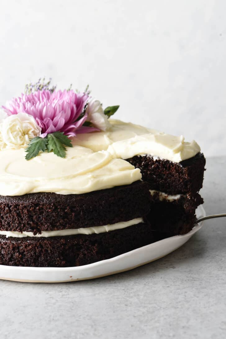 A cake serving sliding a piece of chocolate cake with cream cheese frosting away from the rest of the cake. The cake is two layers and decorated with pink and white flowers.