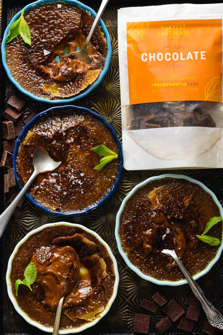 Shallow ramekins of brown custard with torched sugar tops, on a rustic baking pan, with a bag of Harvest Chocolate.