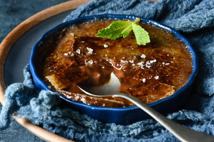 A shallow dark blue ramekin filled with chocolate creme brulee, garnished with a mint sprig and sea salt flakes, with a spoon digging into it.