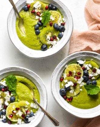 Three fruit smoothie bowls topped with blueberries, pomegranate seeds, coconut, kiwi and a green leaf.