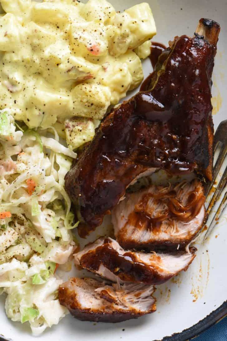 A country style rib with some pieces of meat cut off of it, on a plate with potato salad and coleslaw.