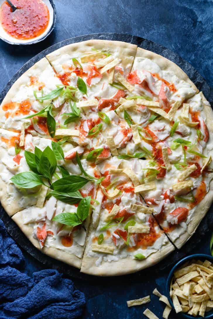 Crab rangoon pizza topped with sweet chili sauce, Thai basil and wonton strips, on blue background.