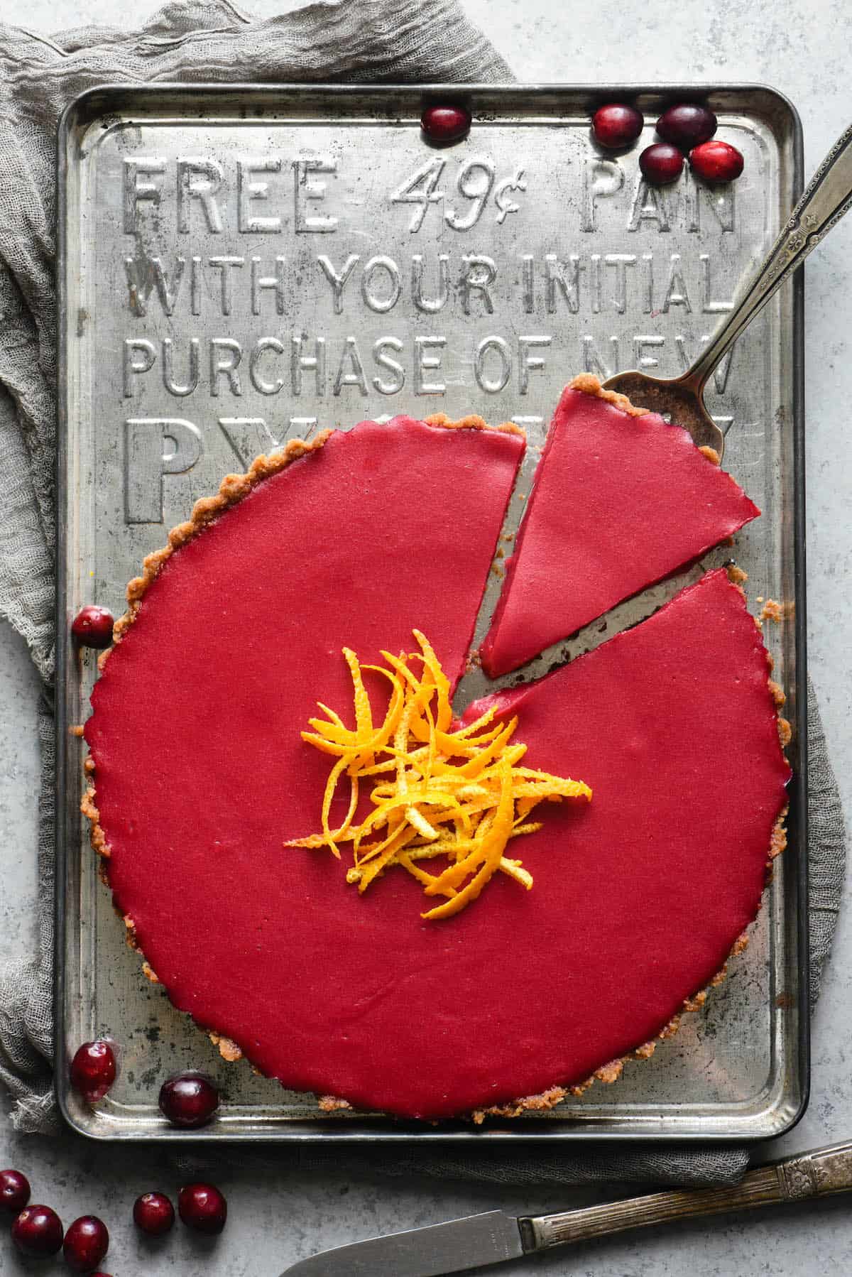 Magenta hued tart on vintage baking sheet. One piece of tart is being lifted out with a pie server. Fresh cranberries garnish scene.