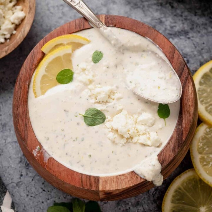 A small wooden bowl filled with creamy feta dressing, garnished with fresh lemon wedges and oregano leaves, with a spoon digging into it.