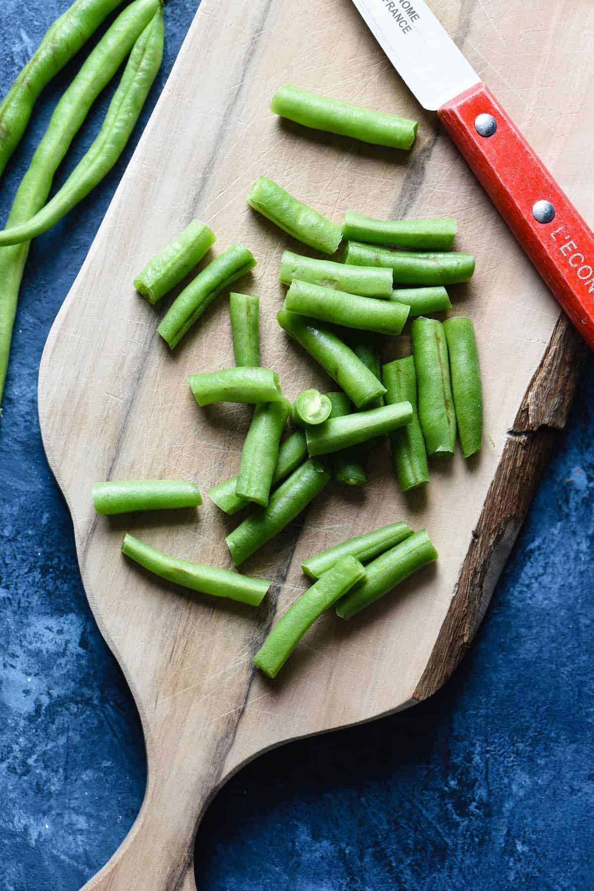 A wooden cutting board and red-handled paring knife, with chopped fresh green beans.