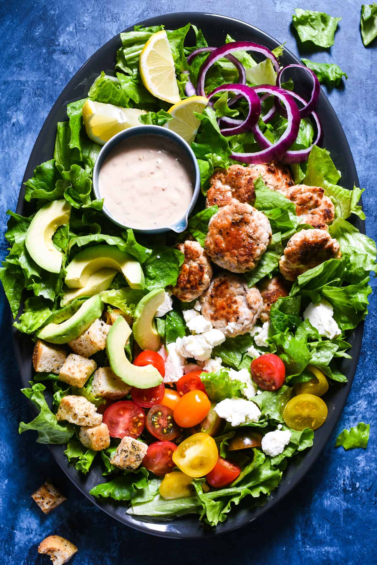 Large platter filled with lettuce, small turkey patties, avocado, croutons, feta cheese, tomatoes and a creamy dressing.