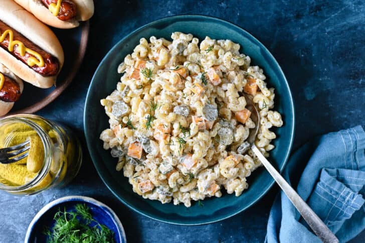 A blue bowl filled with dill pickle pasta salad made with macaroni, fresh dill and cubes of cheddar cheese, with hot dogs alongside.