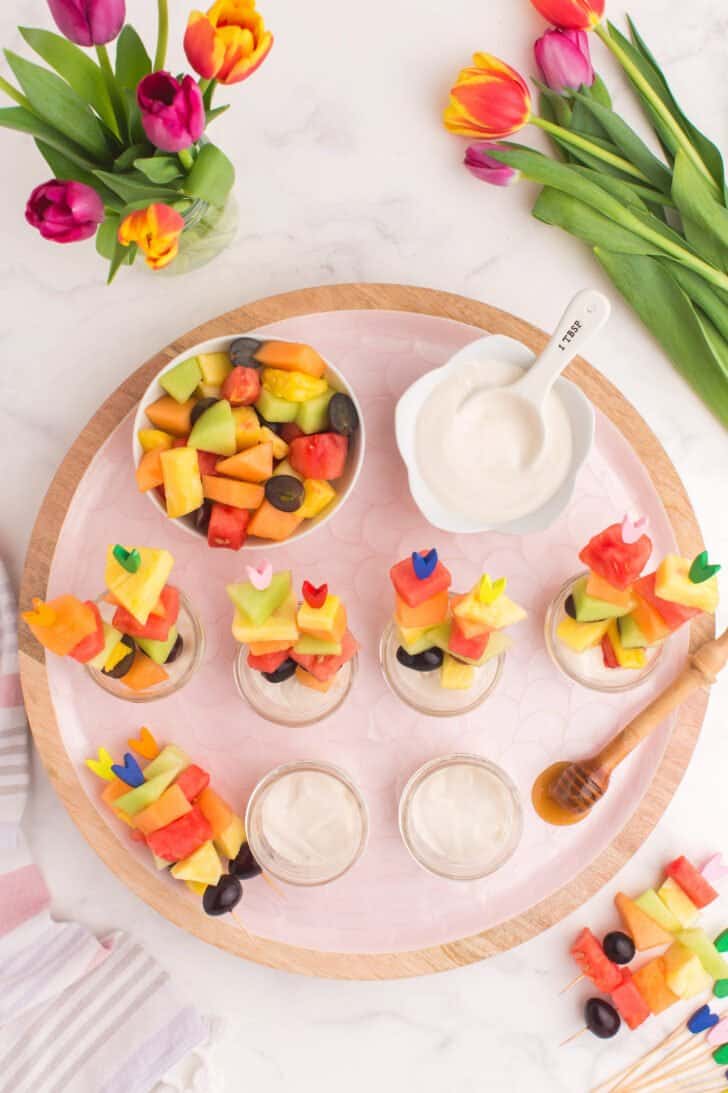 Small jars filled with cottage cheese dip and Easter fruit salad skewers on a pink tray.