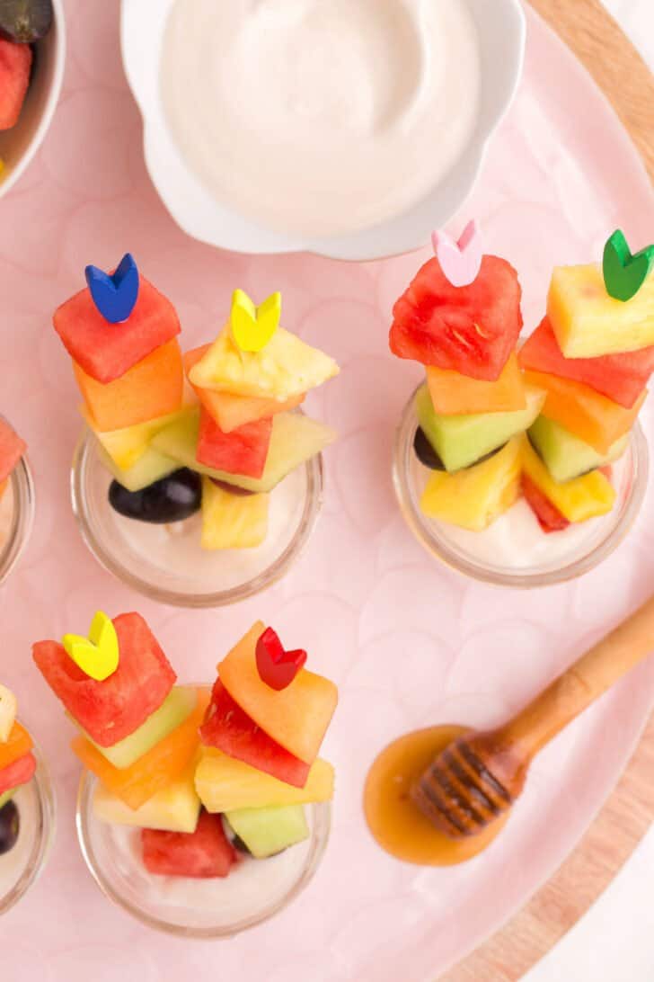 Small jars filled with cottage cheese dip and Easter fruit salad skewers on a pink tray.