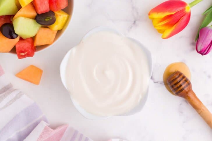 Yogurt fruit dip in a small flower shaped bowl with a honey dipper and fresh fruit nearby.