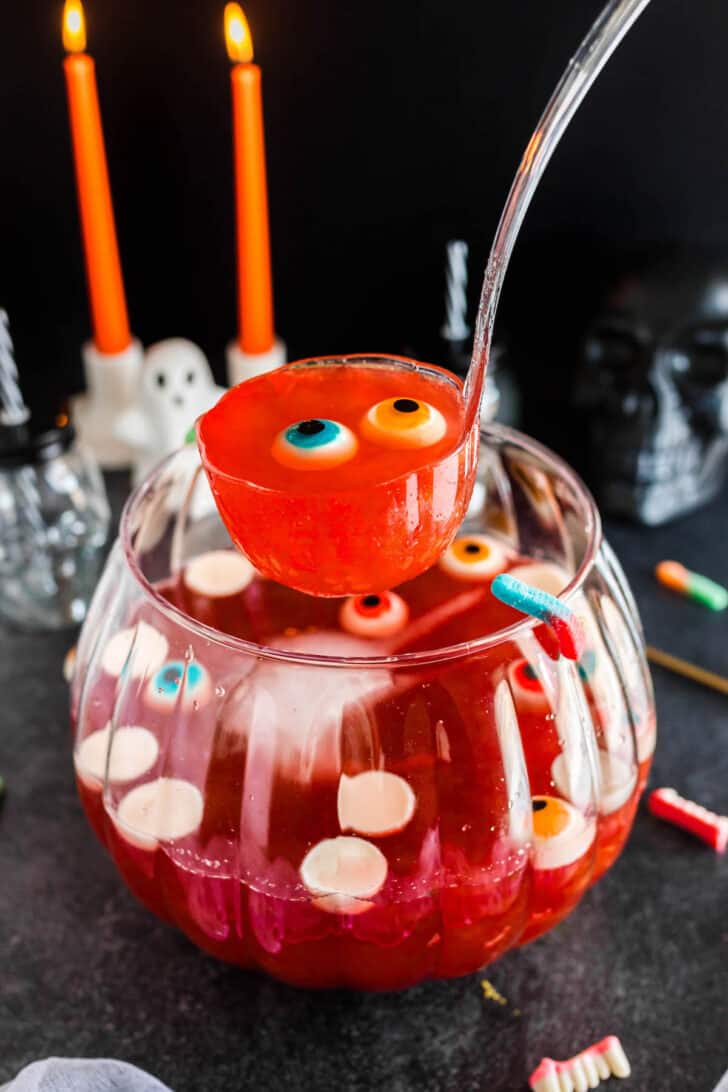 A glass bowl filled with red Halloween punch and gummy eyeballs, with a ladle lifting a serving out.