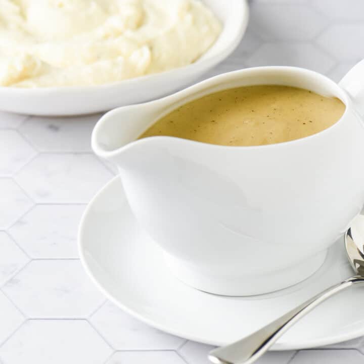 White ceramic gravy boat filled with homemade gravy, with bowl of mashed potatoes in background.