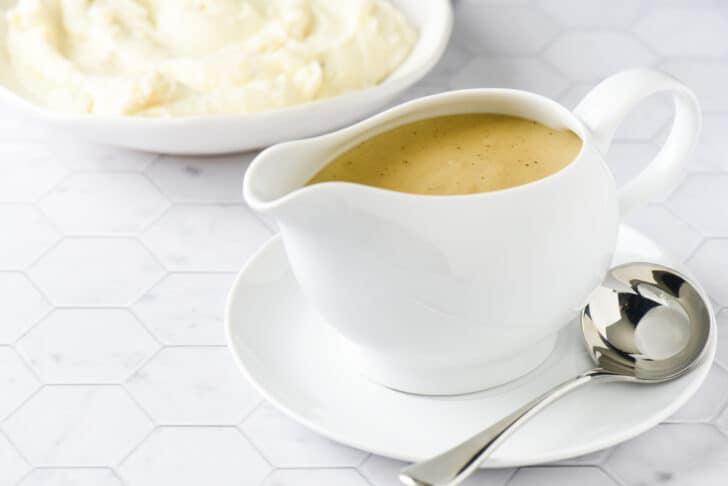 White ceramic gravy boat filled with homemade gravy, with bowl of mashed potatoes in background.