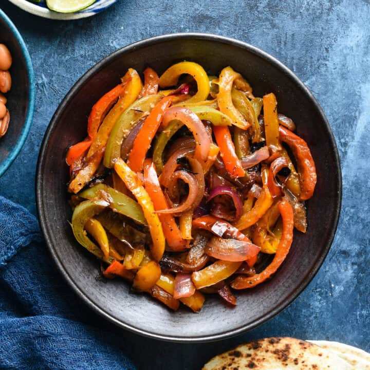 Black bowl filled with cooked fajita veggies on a blue background.