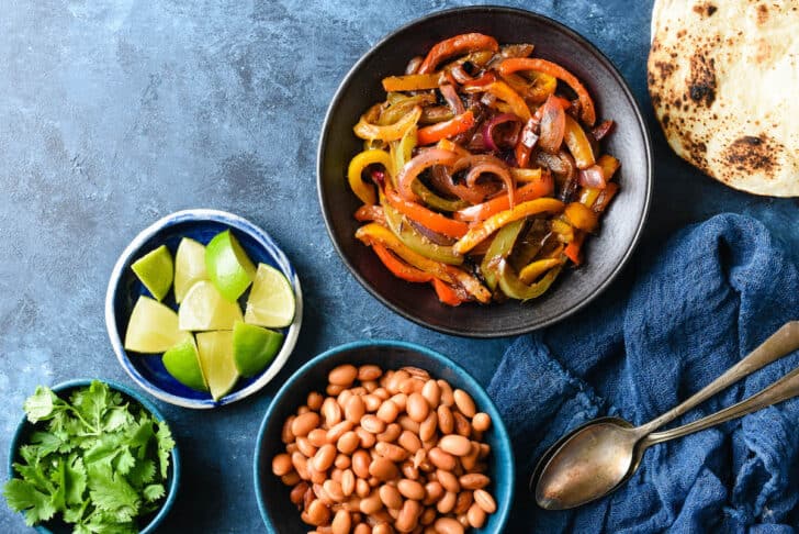 Bowls of sauteed fajitas veggies recipe, pinto beans, lime wedges and cilantro on blue background with charred tortillas.