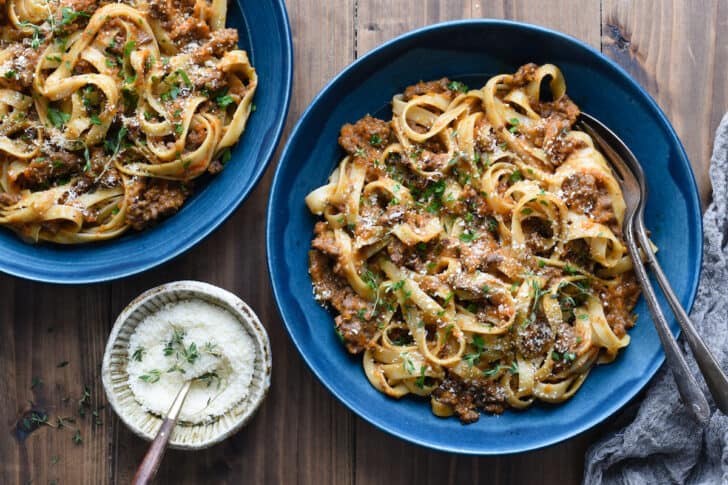 Two blue ceramic bowls filled with fettuccine bolognese on a wooden tabletop, along with a smaller bowl of Parmesan cheese.