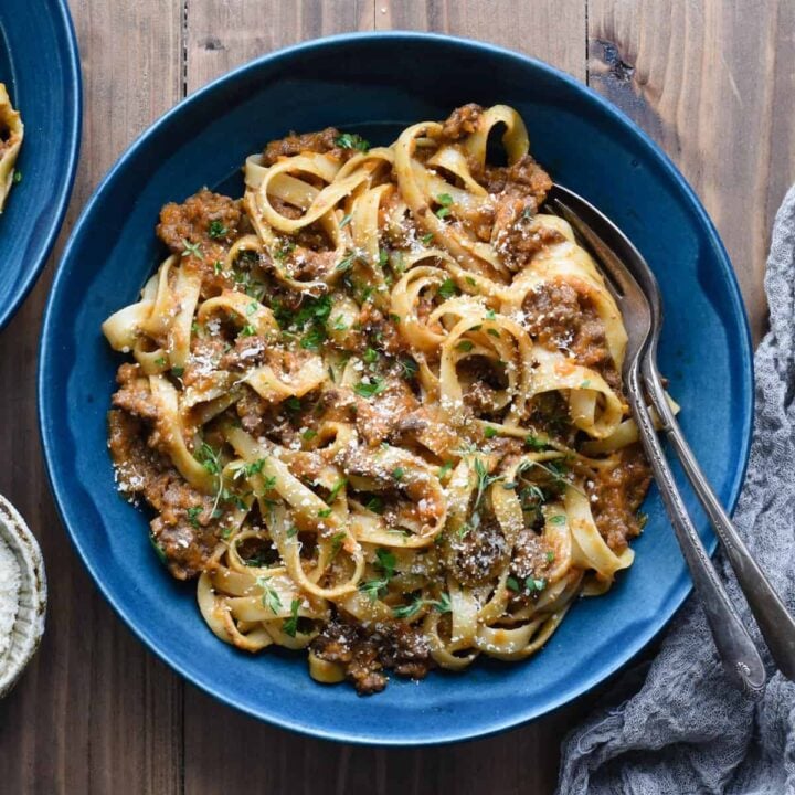 A blue ceramic bowl filled with fettuccine bolognese, with a fork and spoon resting in the bowl.