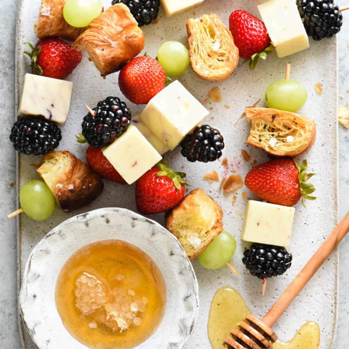 Skewers of fresh berries, white cheese and croissant pieces, with a small bowl of honey.