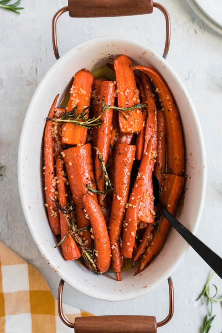 White oval ceramic baking dish with wooden handles filled with garlic honey roasted carrots garnished with rosemary.