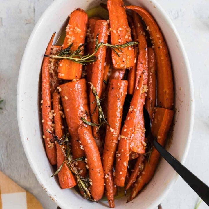 White oval ceramic baking dish with wooden handles filled with garlic honey roasted carrots garnished with rosemary.