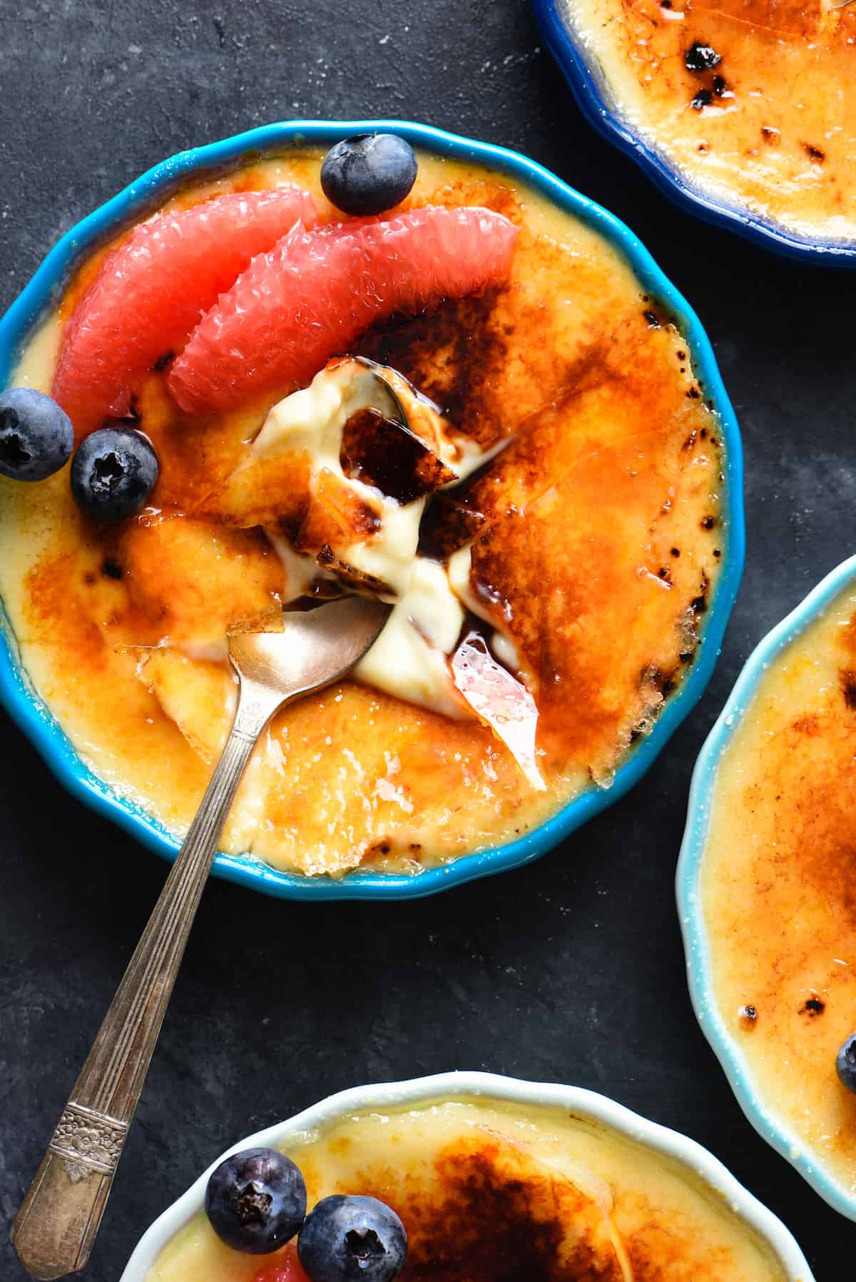 Grapefruit creme brulee in ramekins, garnished with grapefruit segments and blueberries, on a dark surface, with a spoons digging into them.