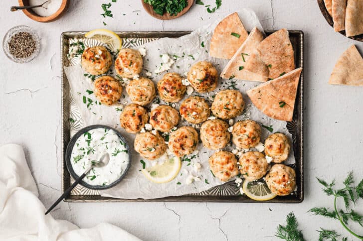 Mediterranean meatballs on a rimmed baking pan with pita wedges, lemon slices and yogurt sauce.