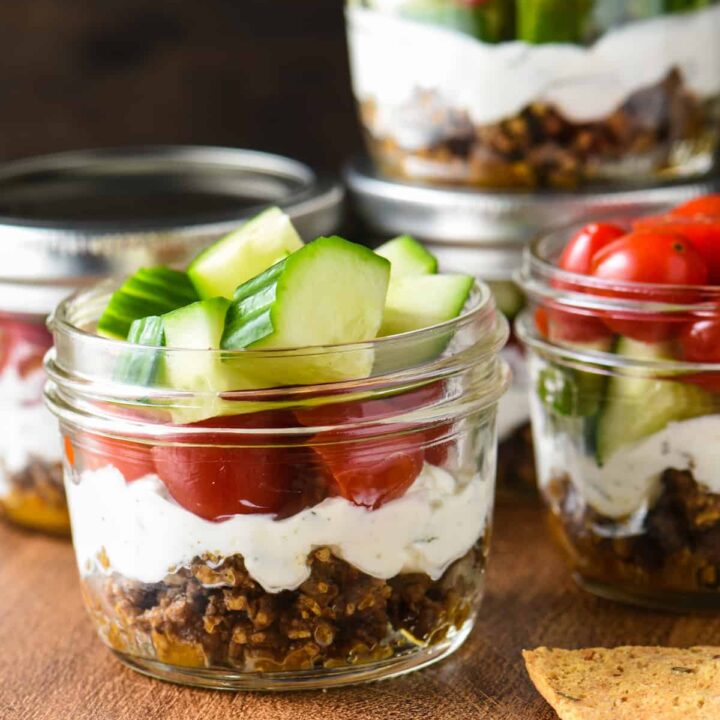 Pint sized mason jars filled with Greek layer dip, including ground lamb, yogurt, cucumbers and tomatoes.