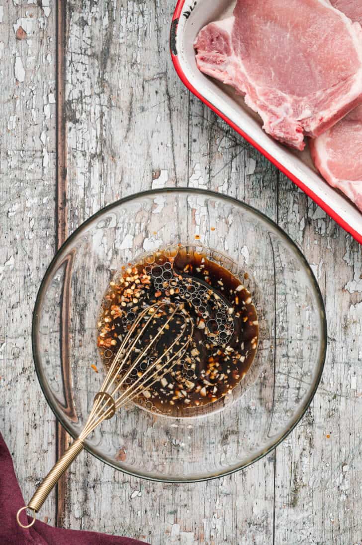 A glass bowl with a metal whisk in a dark liquid mixture with chopped garlic in it.